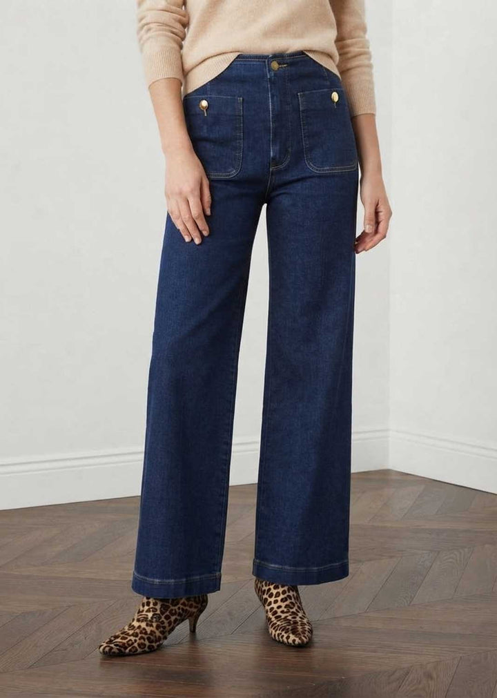 A person wearing madeline dark blue wide-leg cropped jeans with gold-tone button and rivet details, paired with a beige sweater and leopard-print pointed-toe heels, standing in a room with a light wall and wood floor.
