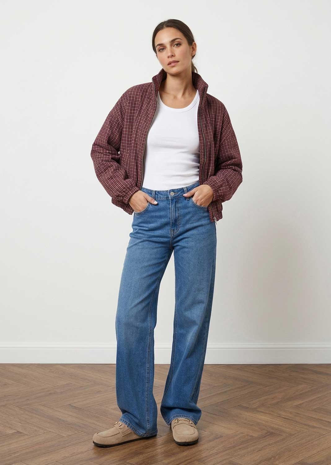 A woman stands in a studio wearing a burgundy plaid zip-up jacket over a white tank top, blue wide-leg jeans, and beige sneakers against a white wall and wood floor.