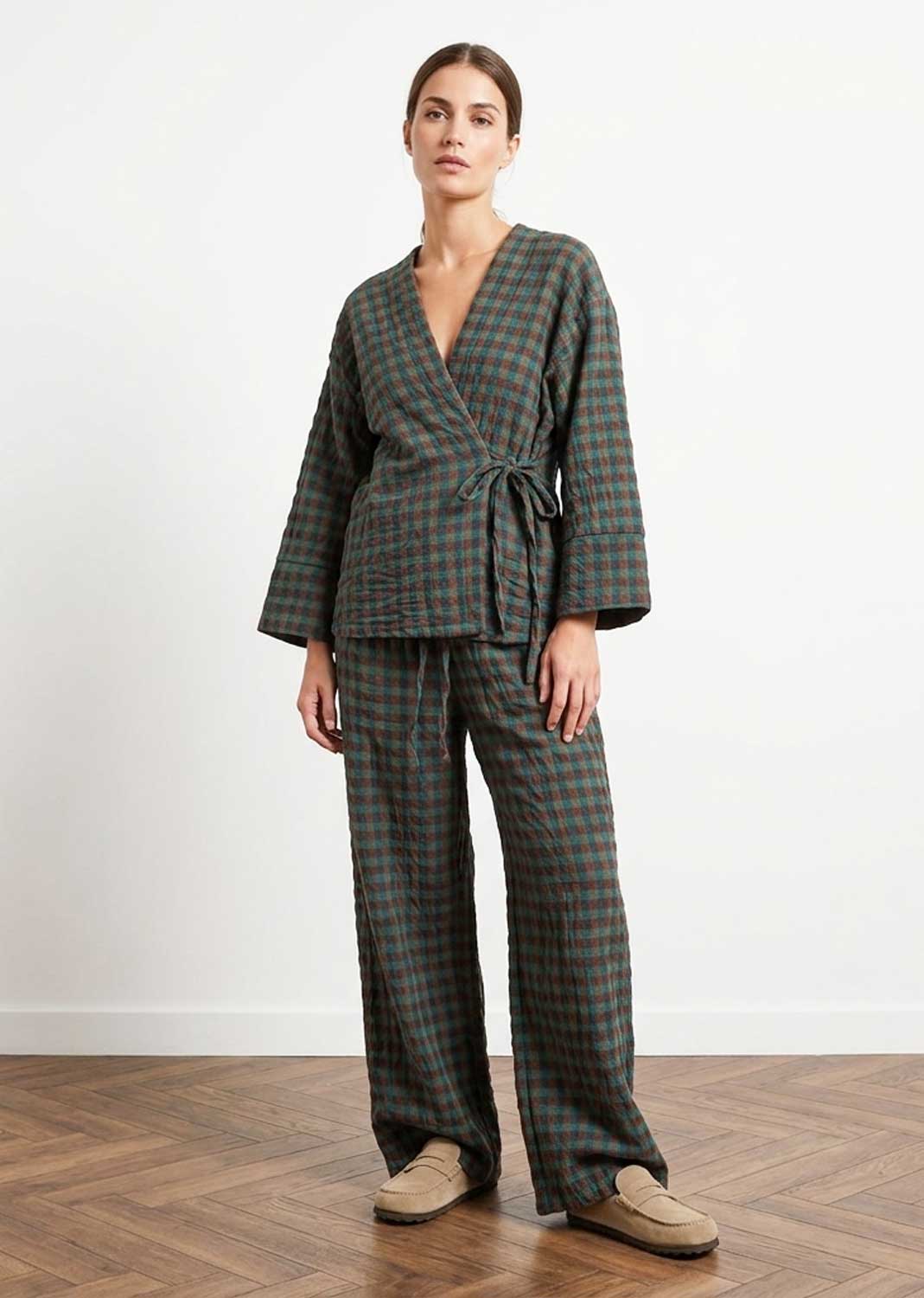 A woman stands in a studio wearing a matching plaid wrap robe and wide-leg trousers set in dark green, brown, and beige tones, paired with tan slip-on shoes against a white wall and wood floor.