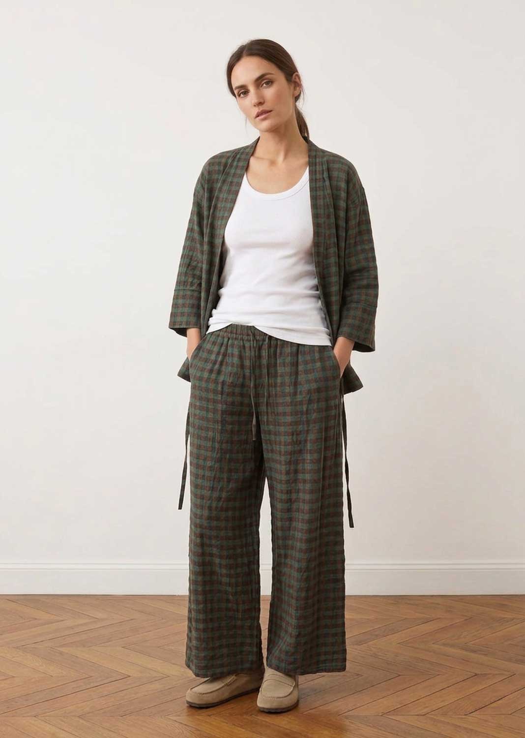 A woman stands in a studio wearing a matching plaid jacket and wide-leg trousers set in dark green and brown tones, layered over a white tank top and beige shoes.