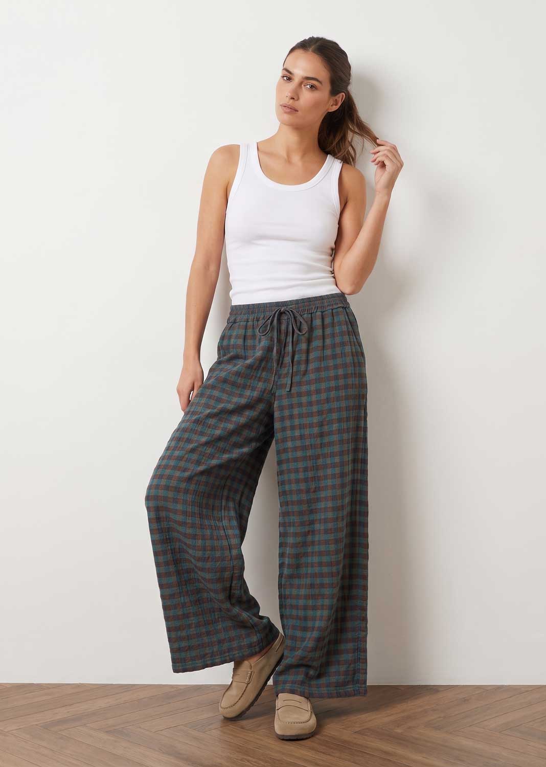 A woman stands against a plain off-white wall wearing wide-leg plaid trousers in dark green and brown, paired with a white tank top and beige slip-on shoes.