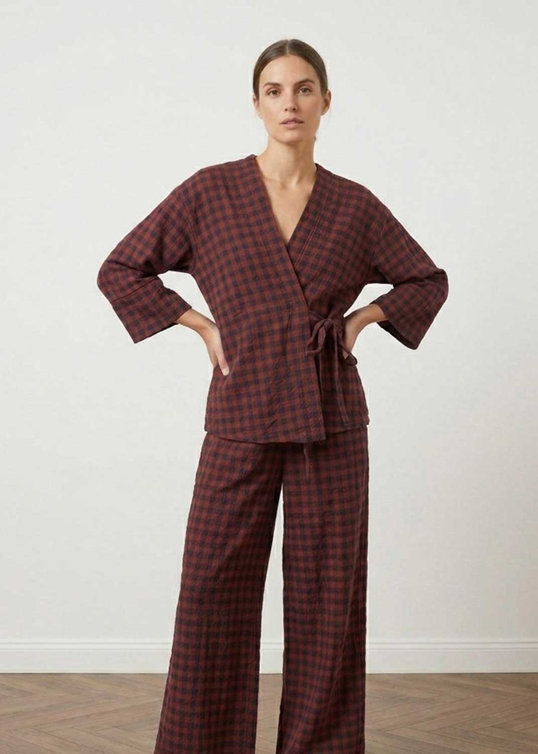 A woman modeling a matching two-piece set with a dark red and black checkered pattern, featuring a wrap-style robe top with long sleeves and wide-leg pants, set against a plain white wall and light wood floor.