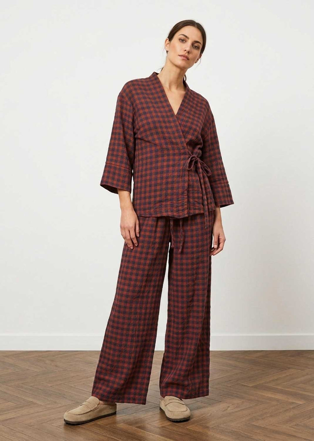 A woman modeling a matching two-piece plaid lounge set consisting of a wrap-style top with three-quarter sleeves and wide-leg pants, in a dark red and black checkered pattern, paired with beige slip-on shoes against a white wall and wood floor.