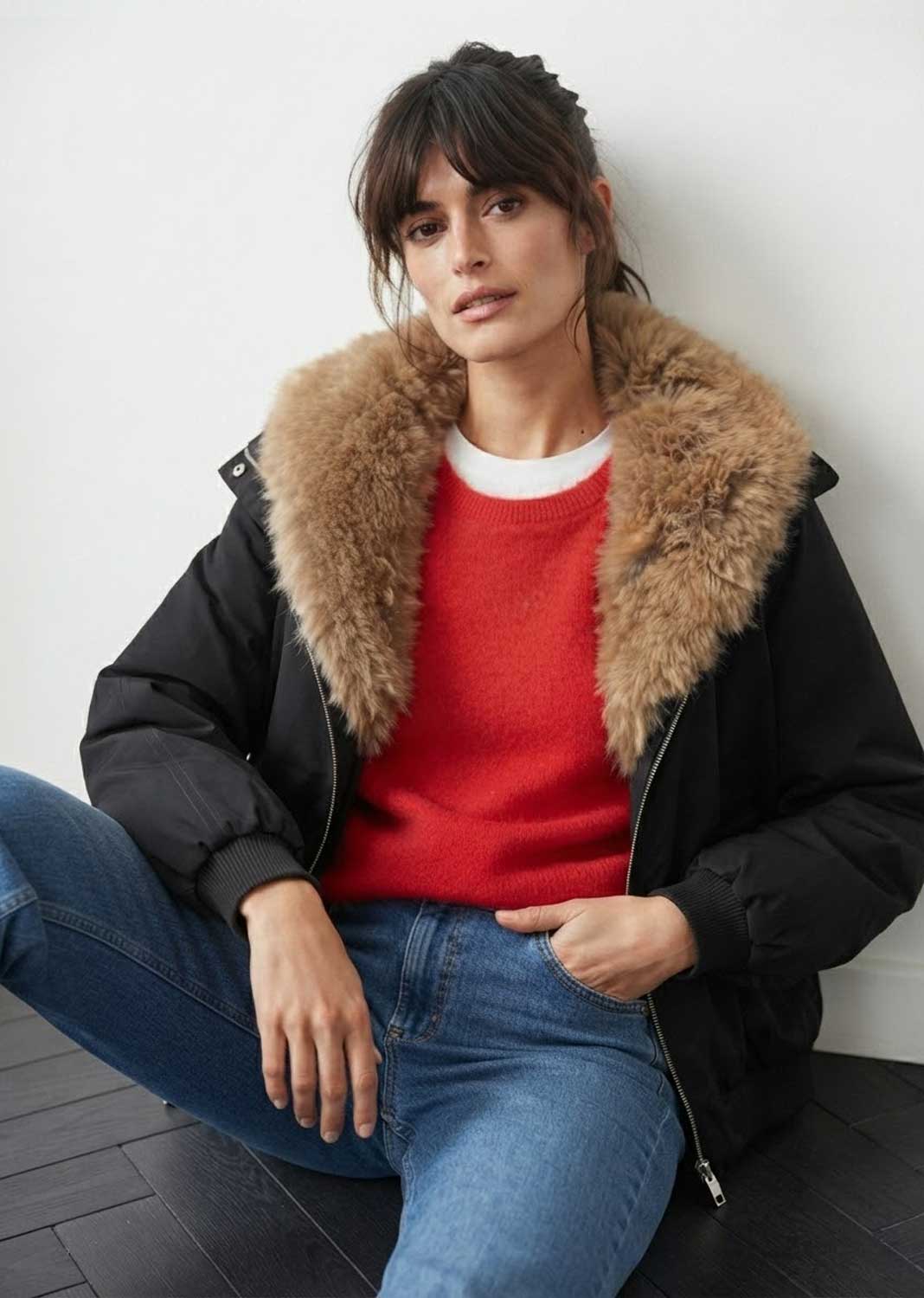 A woman wearing a black puffer jacket with a large tan faux-fur collar, layered over a red knit sweater and blue jeans, sitting against a white wall.