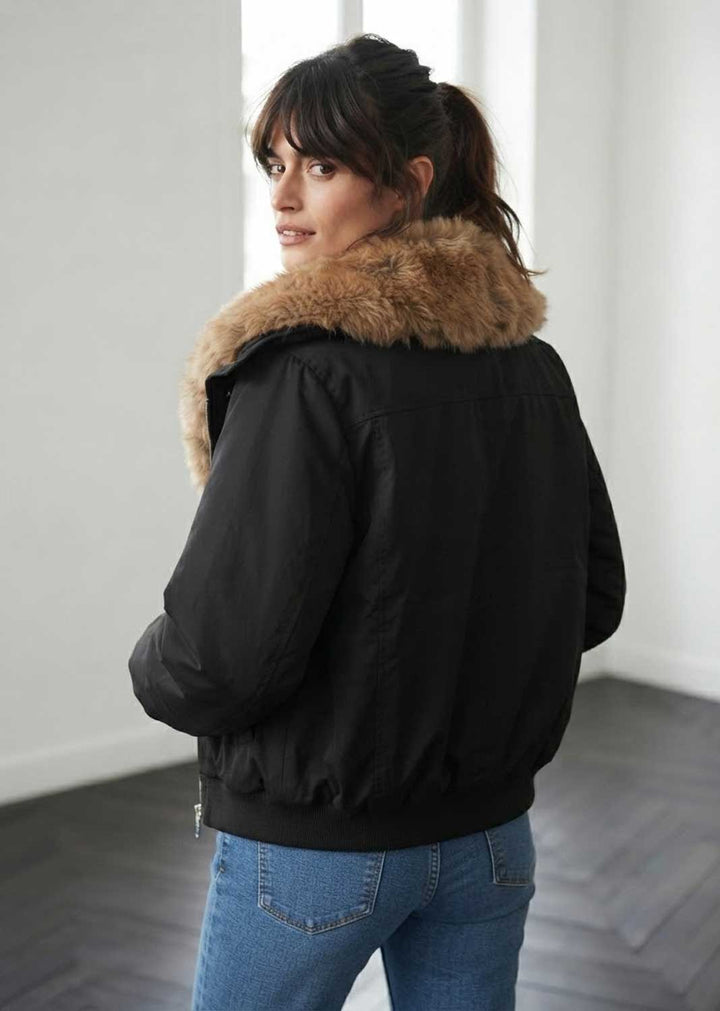 A woman wearing a black bomber jacket with a large tan faux-fur collar, shown from the back in an indoor setting with a white wall and dark wood floor.
