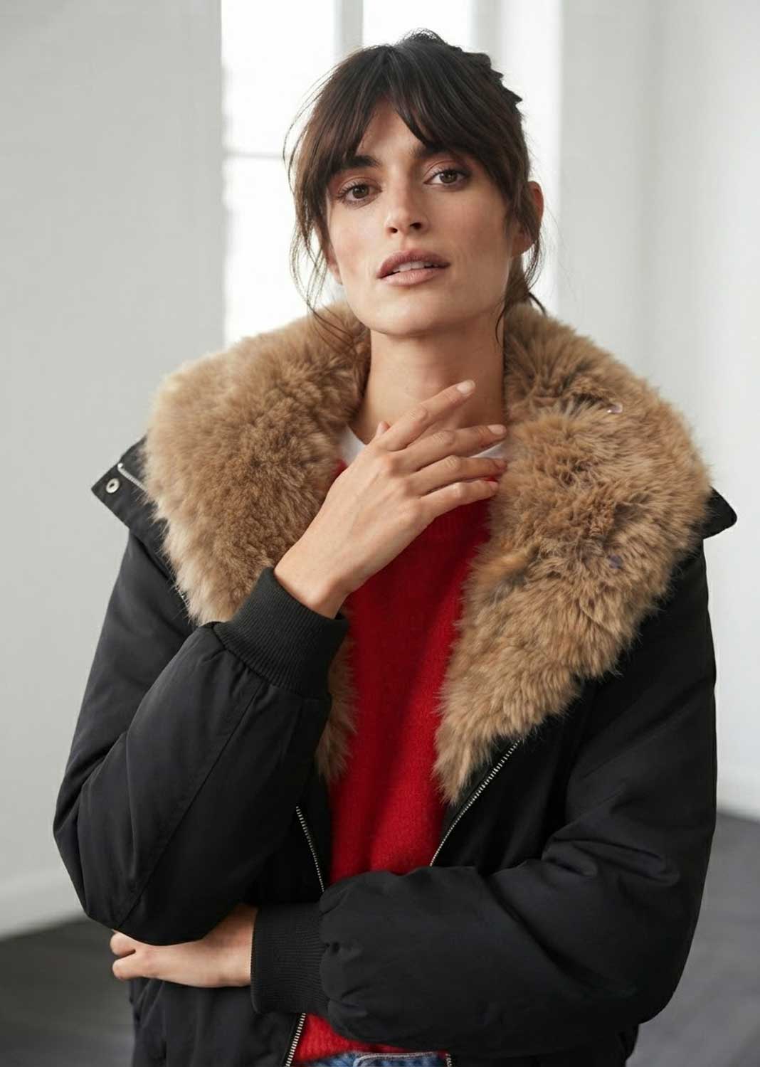 A woman wearing a black puffer jacket with a large tan-brown fur collar over a red sweater, standing in a bright indoor setting.