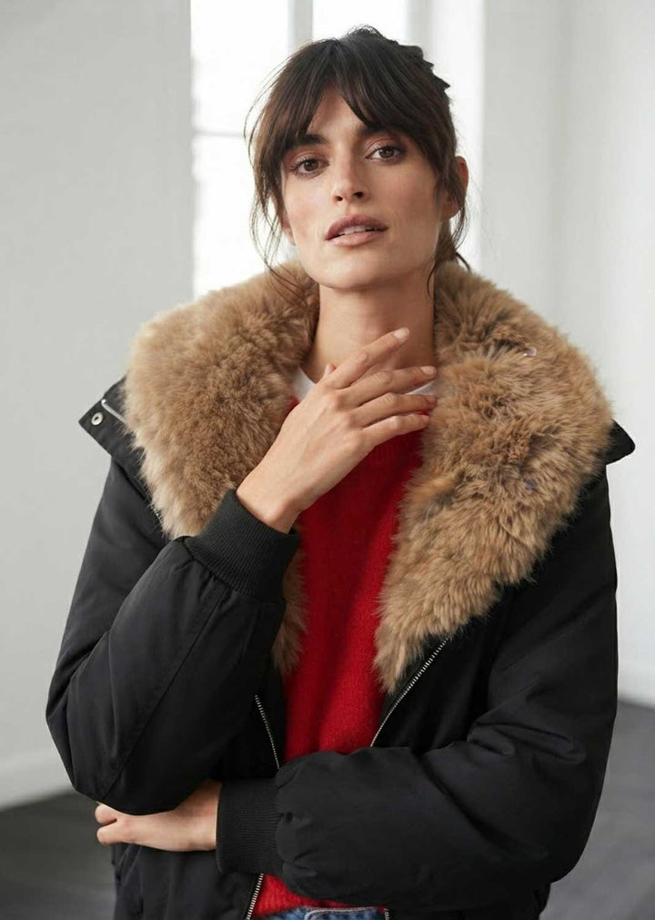 A woman wearing a black puffer jacket with a large tan-brown fur collar over a red sweater, standing in a bright indoor setting.