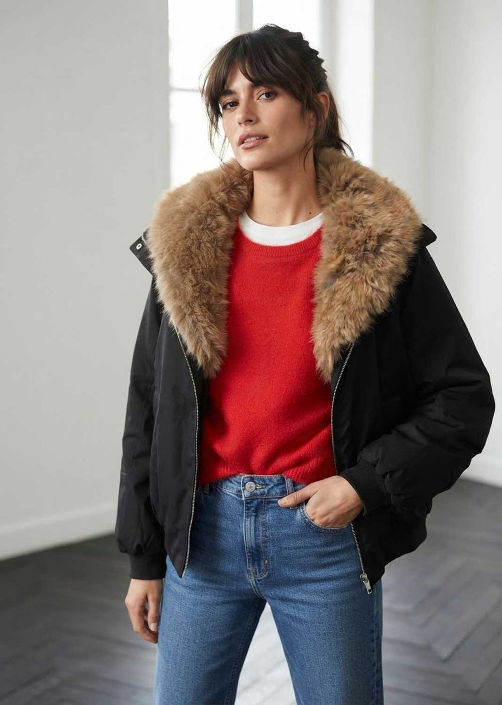 A woman wearing a black puffer jacket with a large brown fur collar, layered over a red knit sweater and blue jeans, standing in a bright indoor setting.