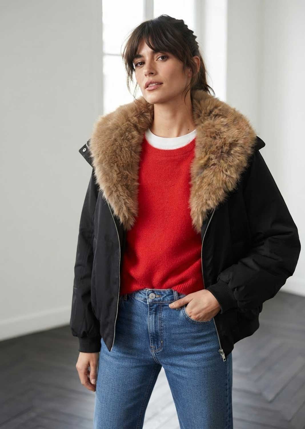 A woman wearing a black puffer jacket with a large brown fur collar, layered over a red knit sweater and blue jeans, standing in a bright indoor setting.
