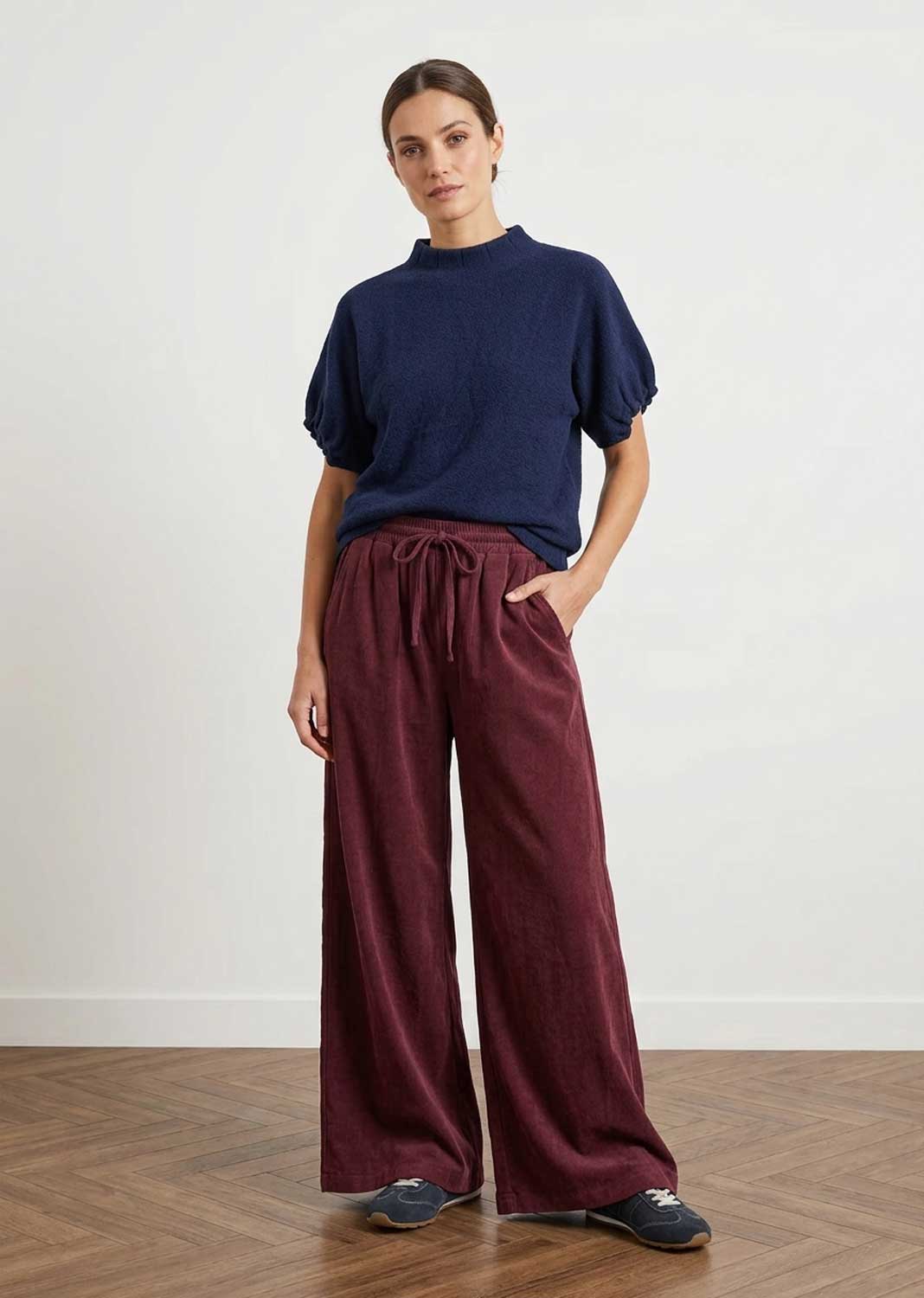 A woman modeling a navy blue short-sleeve mock-neck top and burgundy wide-leg drawstring pants against a white wall and wood floor.