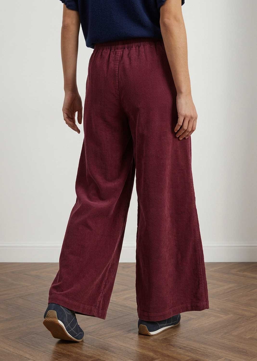 A person shown from the back wearing loose, wide-leg burgundy corduroy trousers, a navy top, and dark sneakers in an indoor room with a light wall and wood floor.