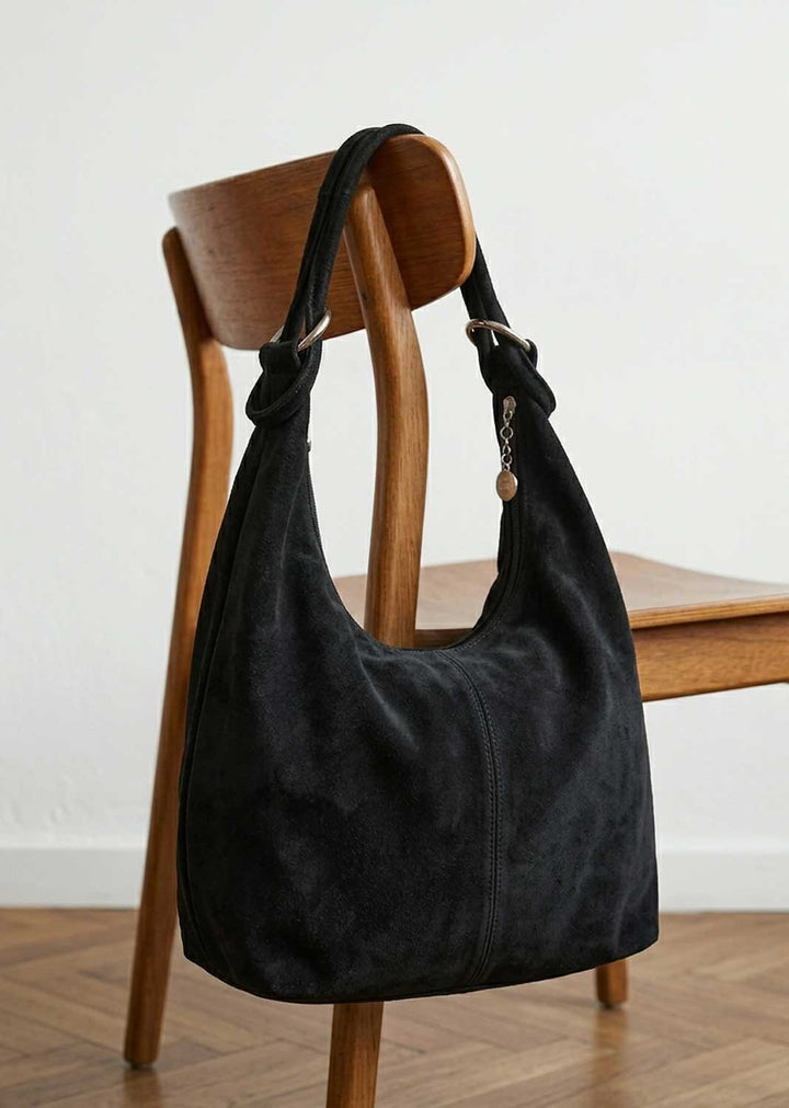 A black suede hobo shoulder bag with a knotted strap detail, draped over a wooden chair in a minimalist room setting.