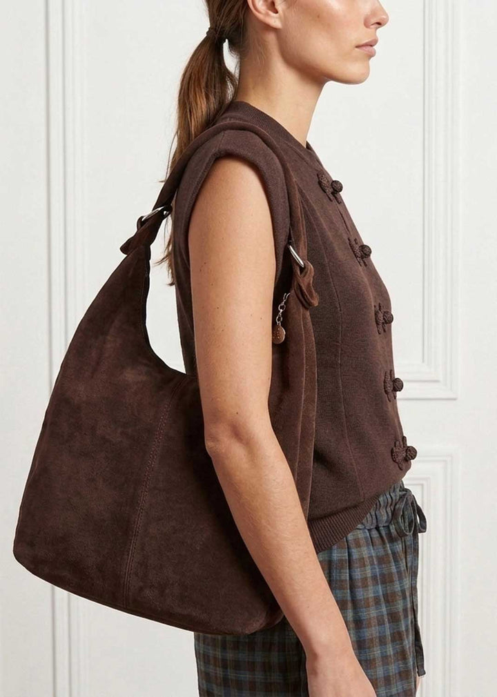A woman wearing a brown suede slouchy shoulder bag over her shoulder, paired with a brown sleeveless button-front top and plaid trousers, set against a white paneled wall.