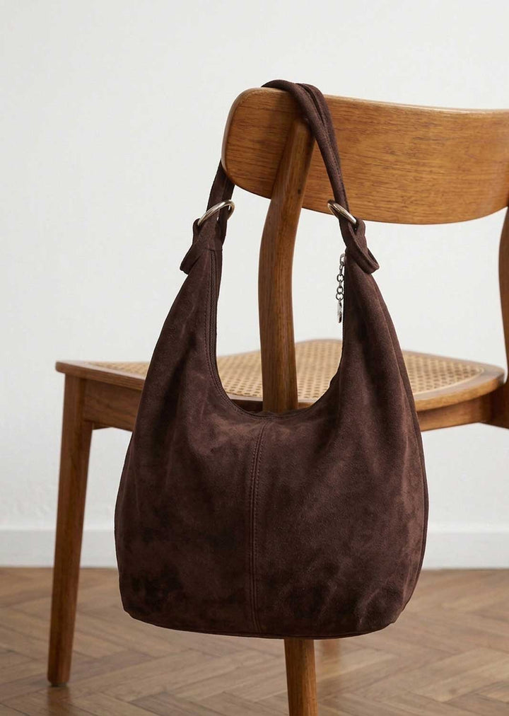 A dark brown suede hobo shoulder bag hanging from the back of a wooden chair in a minimalist room with a light wall and wood floor.