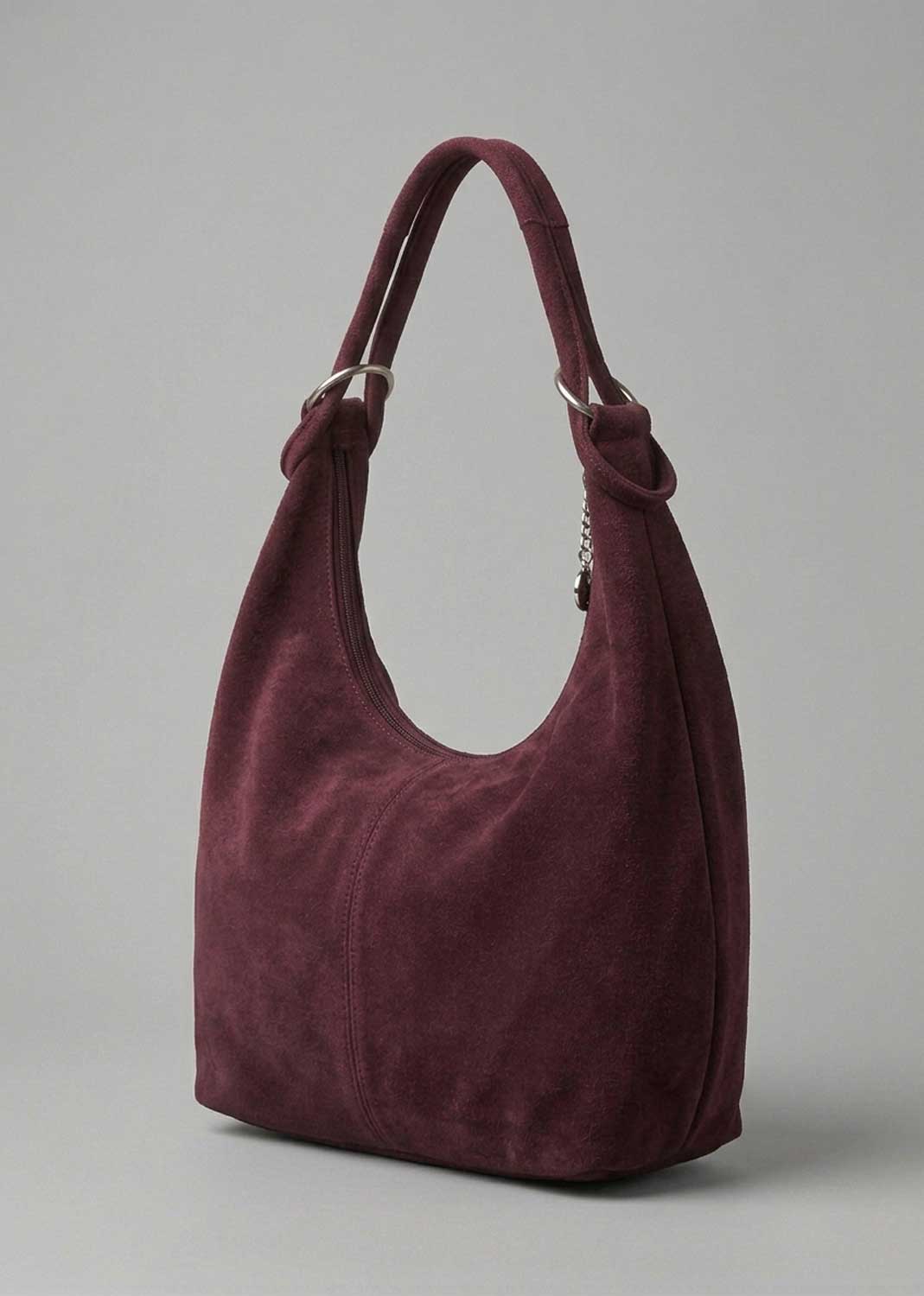 A slouchy hobo shoulder bag in deep burgundy suede, featuring knotted details at the strap attachments and silver-tone hardware, set against a plain gray studio background.