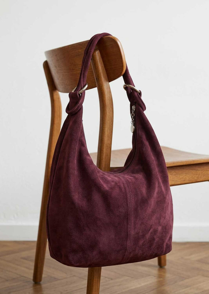 A slouchy suede hobo bag in deep burgundy, draped over a wooden chair in a minimalist room setting.