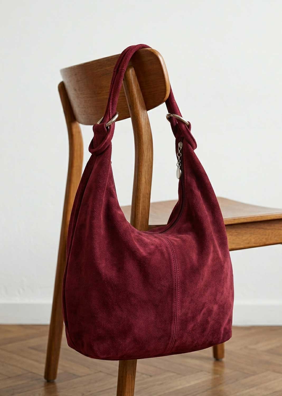 A burgundy suede hobo shoulder bag with a knotted top handle, resting on a wooden chair in a minimalist room setting.