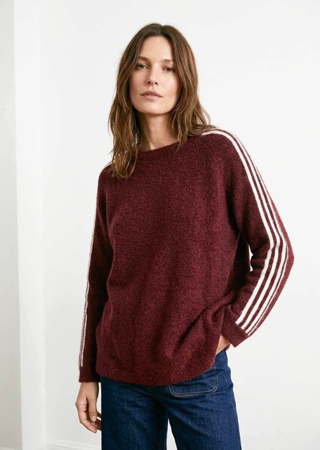 A woman wearing a maroon knit sweater with white stripes down the sleeves, paired with blue jeans, standing against a plain white wall.