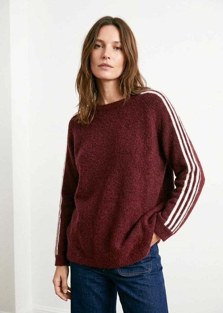 A woman wearing a maroon knit sweater with white stripes down the sleeves, paired with blue jeans, standing against a plain white wall.