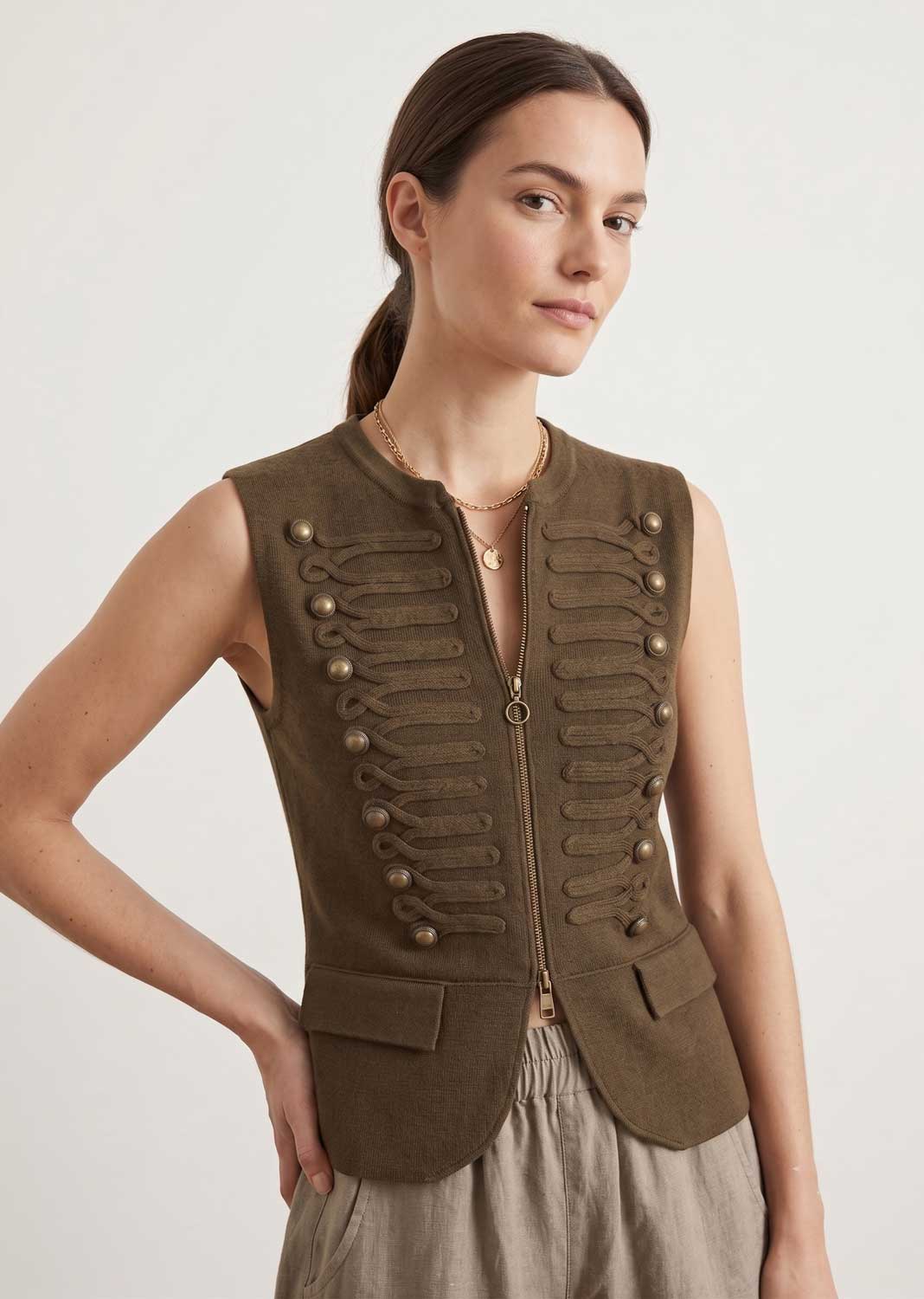 A woman modeling a sleeveless olive-brown military-style vest with decorative button details and a front zipper, paired with light beige trousers, against a plain light gray background.