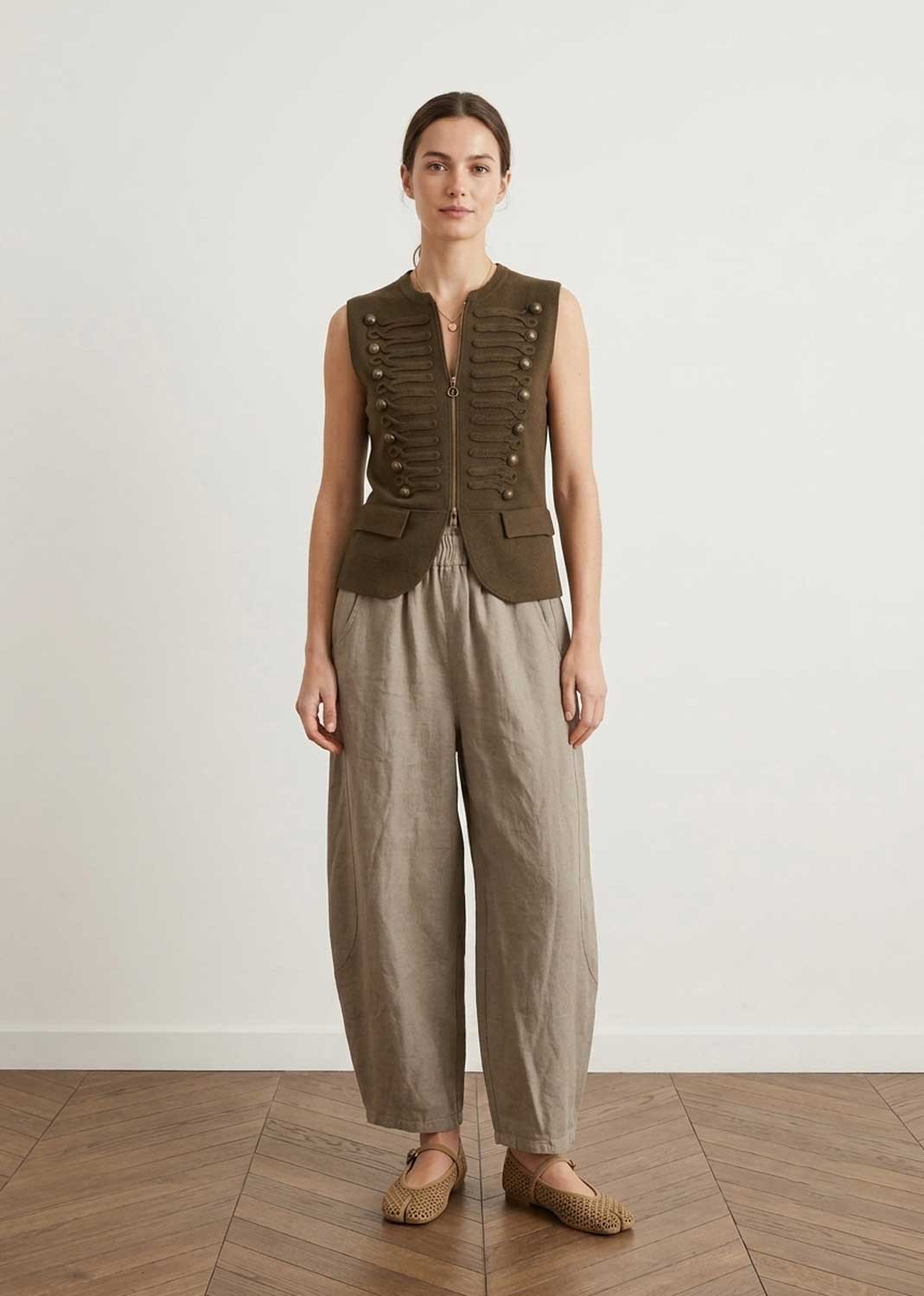 A model stands in a minimalist studio setting wearing a sleeveless olive green military-style vest with gold buttons and beige wide-leg trousers.