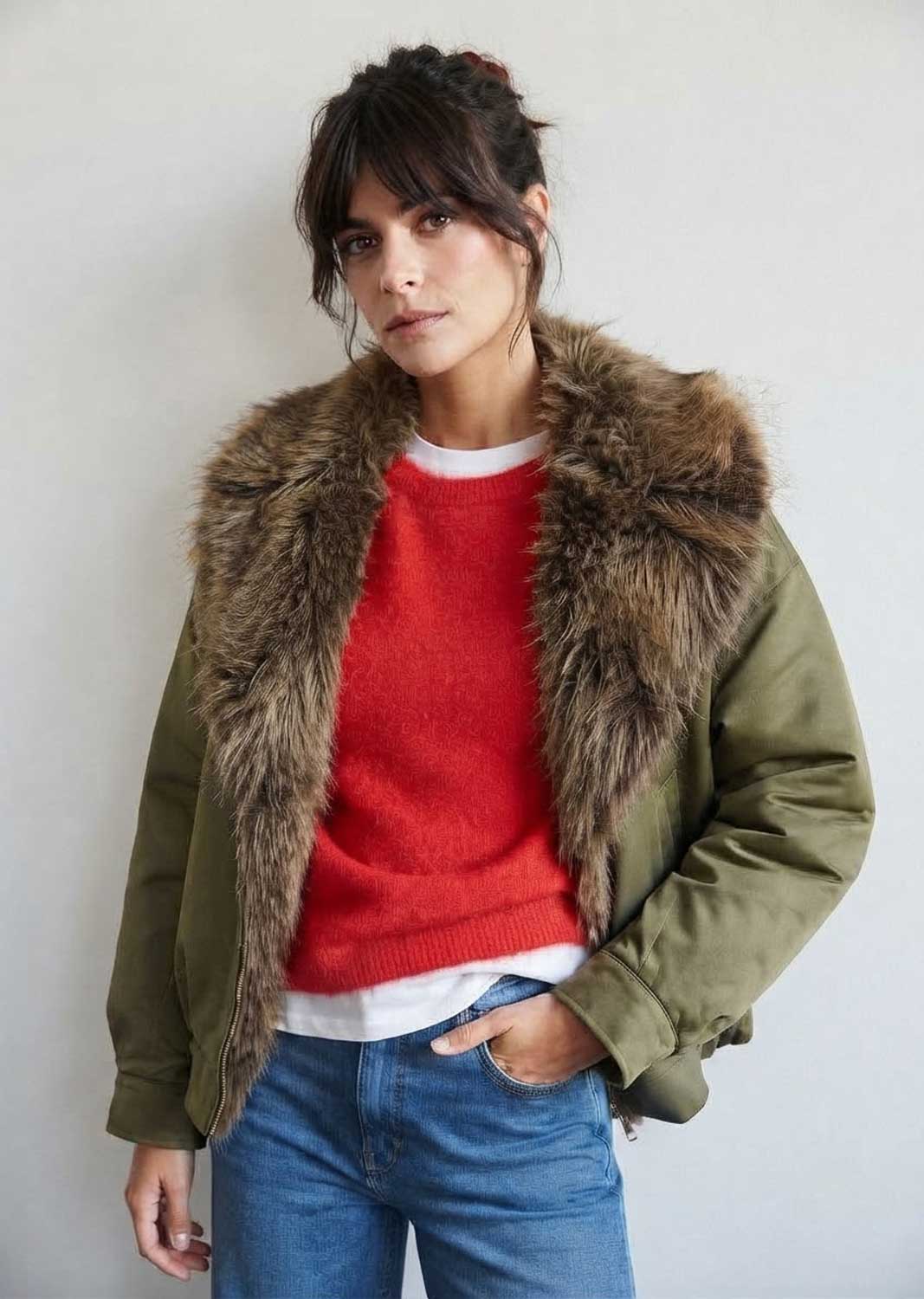A woman wearing an olive green puffer jacket with a large brown faux-fur collar, layered over a red knit sweater and blue jeans,