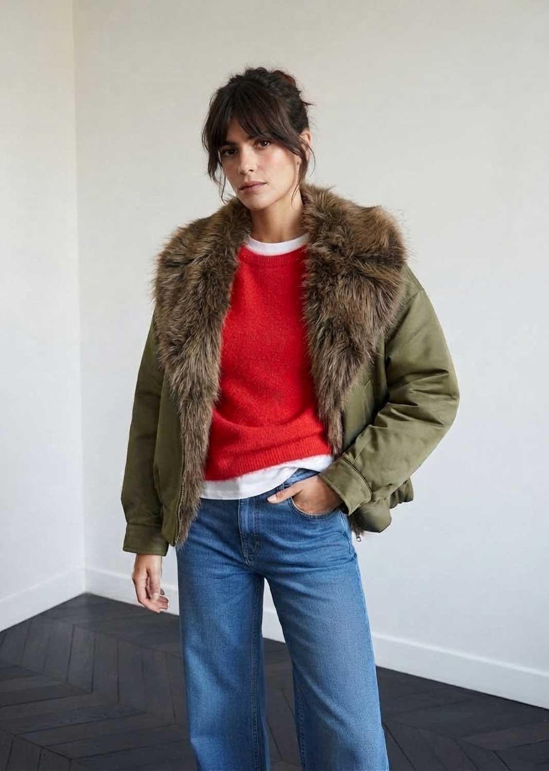 A woman wearing an olive green puffer jacket with a large brown faux-fur collar, layered over a red knit sweater and blue jeans, standing in a minimalist room with a white wall and dark wood floor.