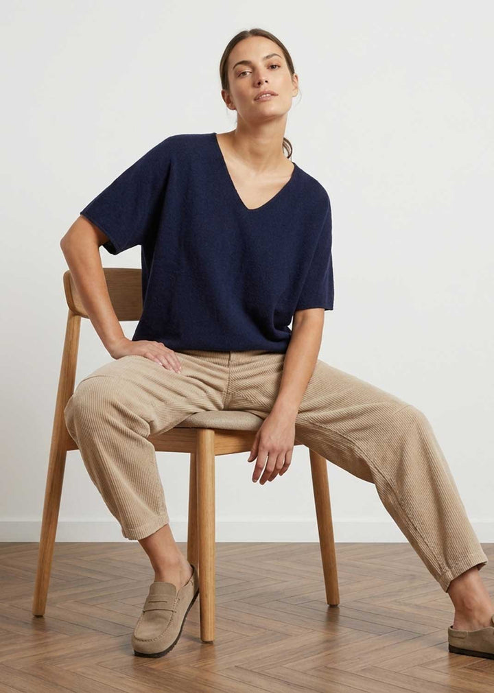 A woman sits on a wooden chair wearing a navy blue short-sleeve V-neck top, beige cropped trousers, and beige loafers in a bright room with a light wall and wood floor.