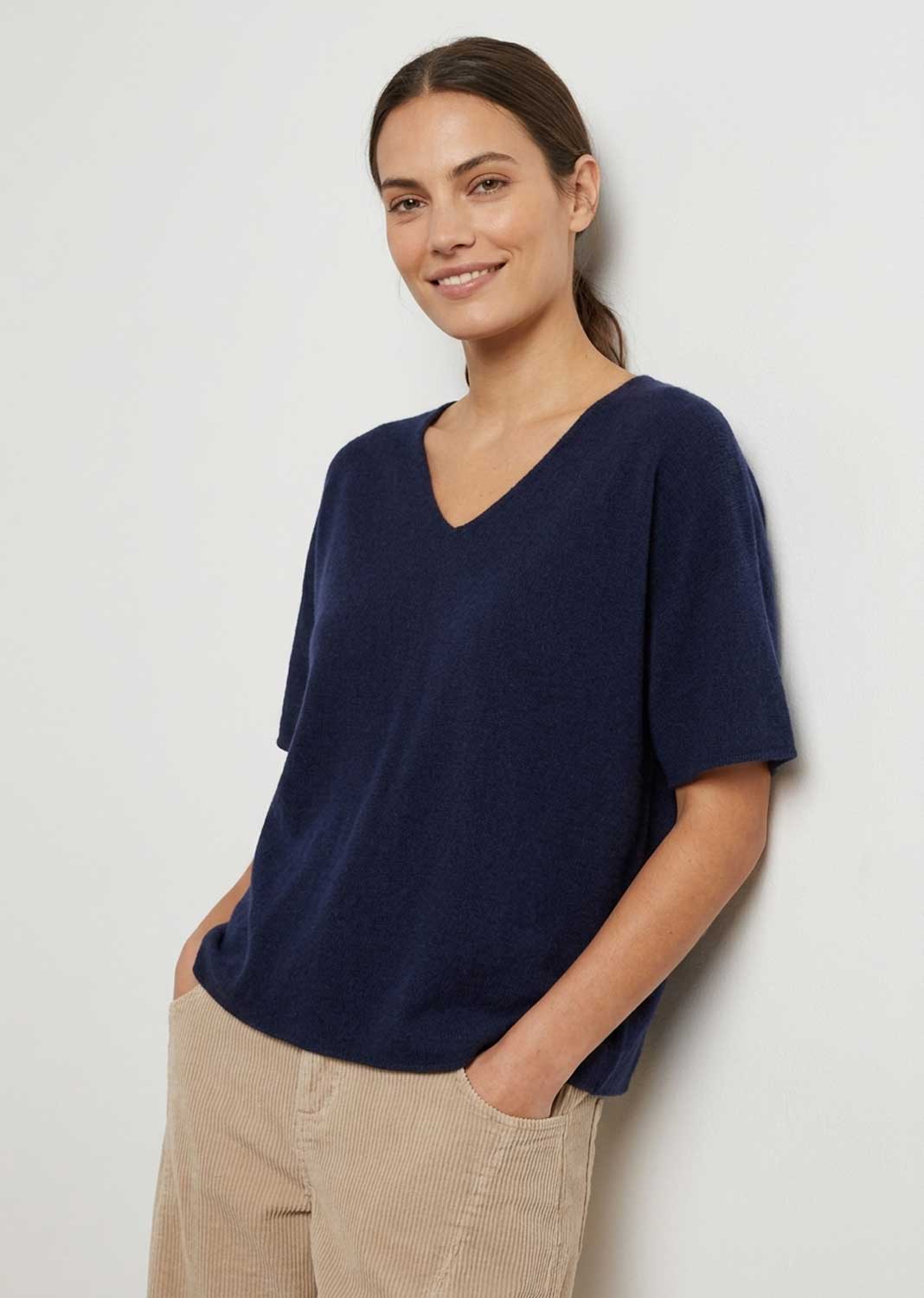 A woman wearing a short-sleeve V-neck knit top in dark navy blue, paired with beige corduroy pants, standing against a plain light gray wall.