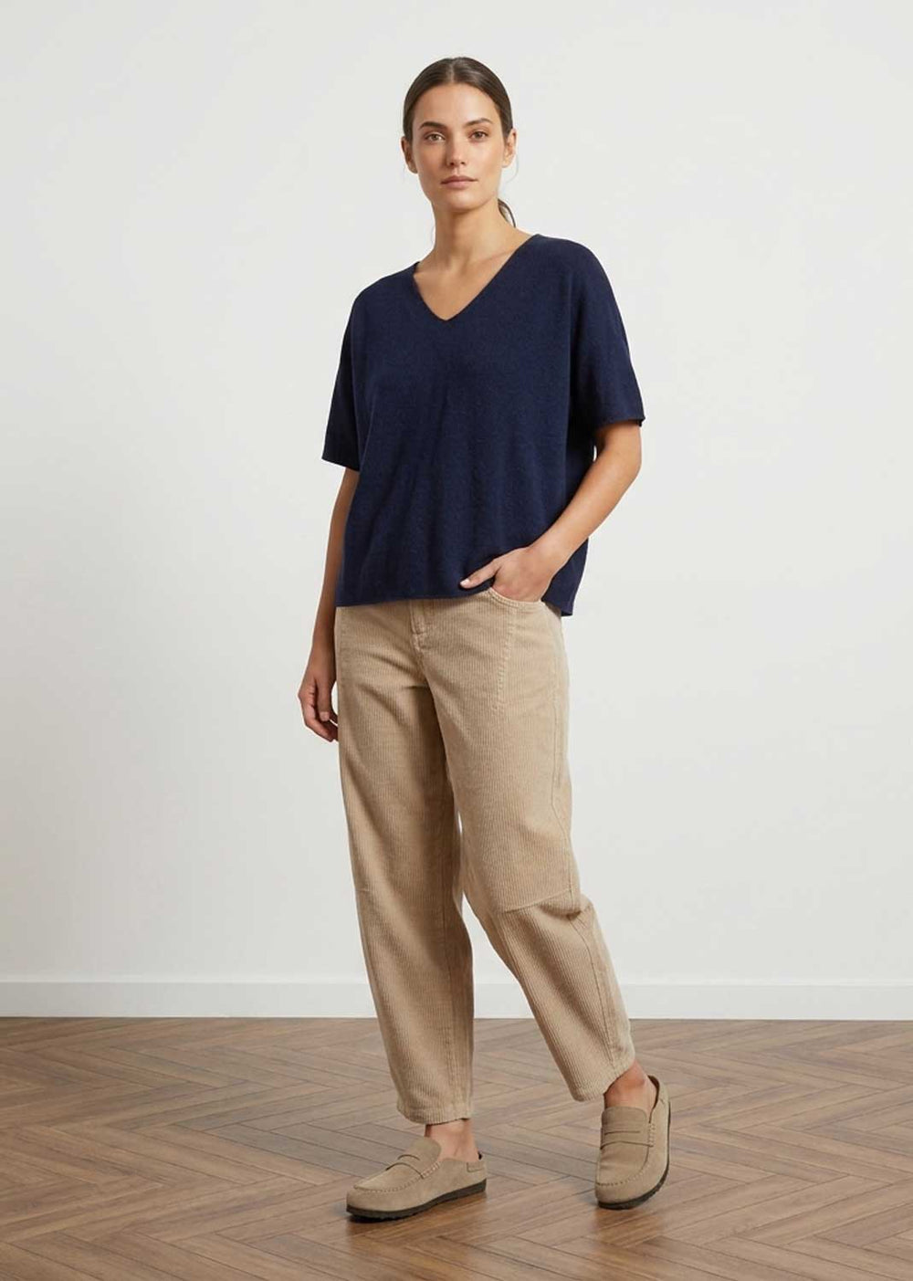 A woman modeling a navy blue short-sleeve V-neck top and beige linen trousers with beige loafers, standing on a wooden parquet floor against a white wall.