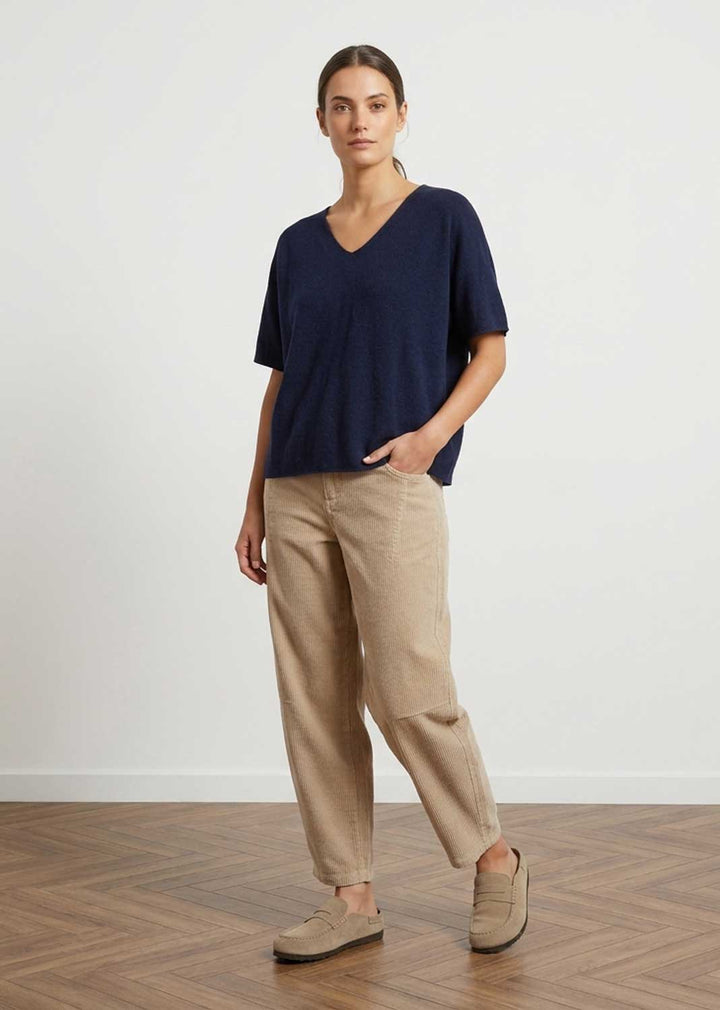 A woman modeling a navy blue short-sleeve V-neck top and beige linen trousers with beige loafers, standing on a wooden parquet floor against a white wall.