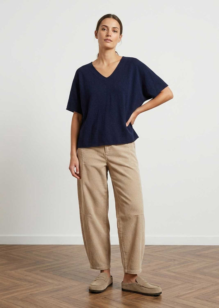 A woman modeling a navy blue short-sleeve V-neck top and beige wide-leg trousers with beige slip-on shoes, standing on a wooden parquet floor against a white wall.