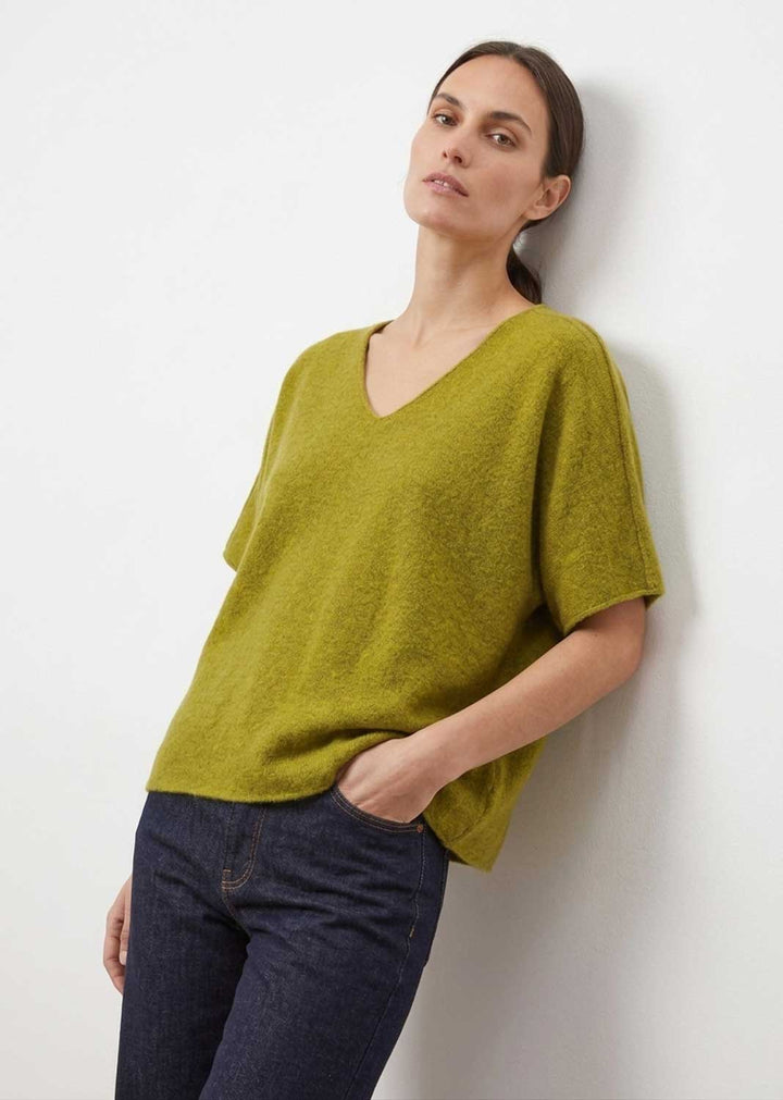 A woman wearing a short-sleeve V-neck knit top in olive green, paired with dark blue jeans, leaning against a plain white wall.