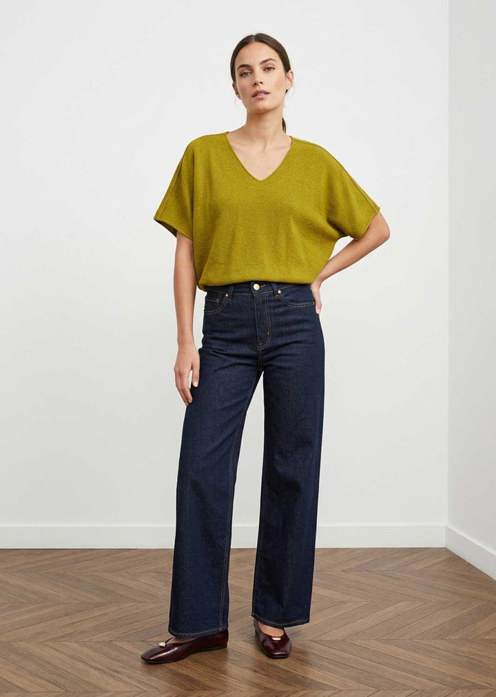 A woman modeling a mustard yellow short-sleeve V-neck knit top and dark blue wide-leg jeans, standing on a wooden floor against a white wall.