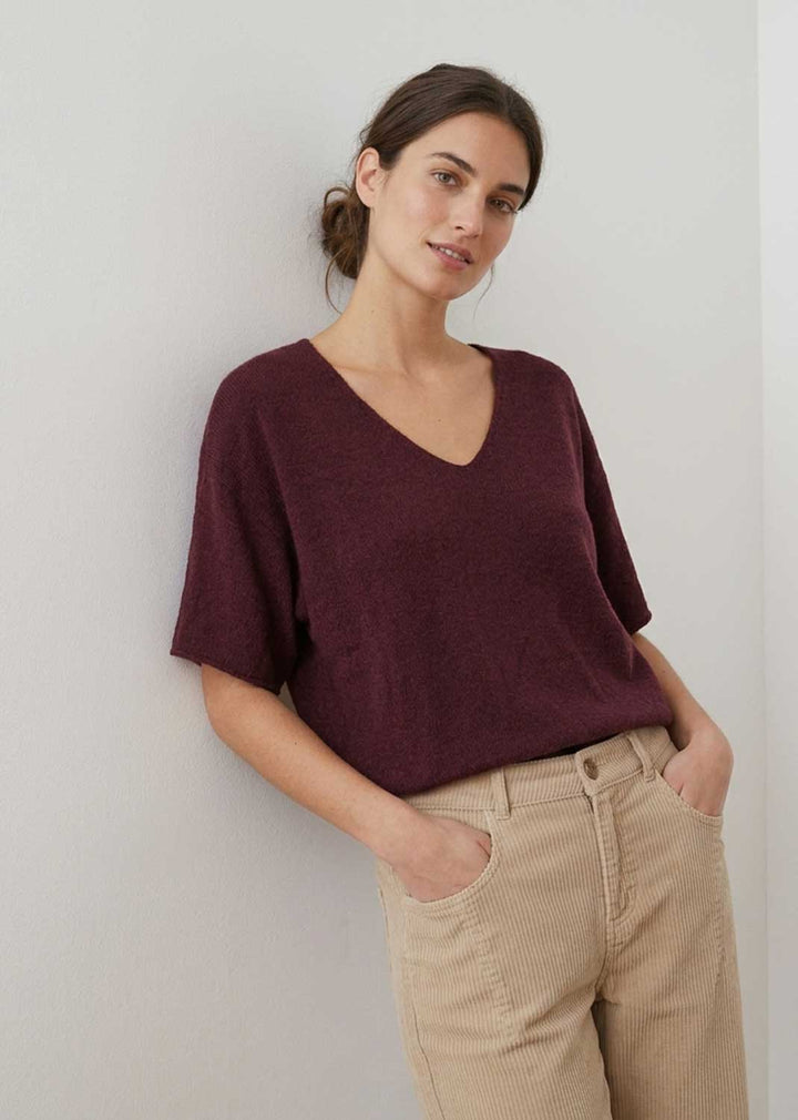 A woman wearing a short-sleeve V-neck knit top in deep burgundy and beige corduroy pants, leaning against a light gray wall.
