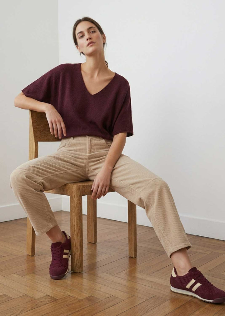 A woman sits on a wooden chair in a minimalist room with a light wall and wood parquet floor, wearing a dark burgundy V-neck short-sleeve top, beige trousers, and burgundy sneakers with white stripes.