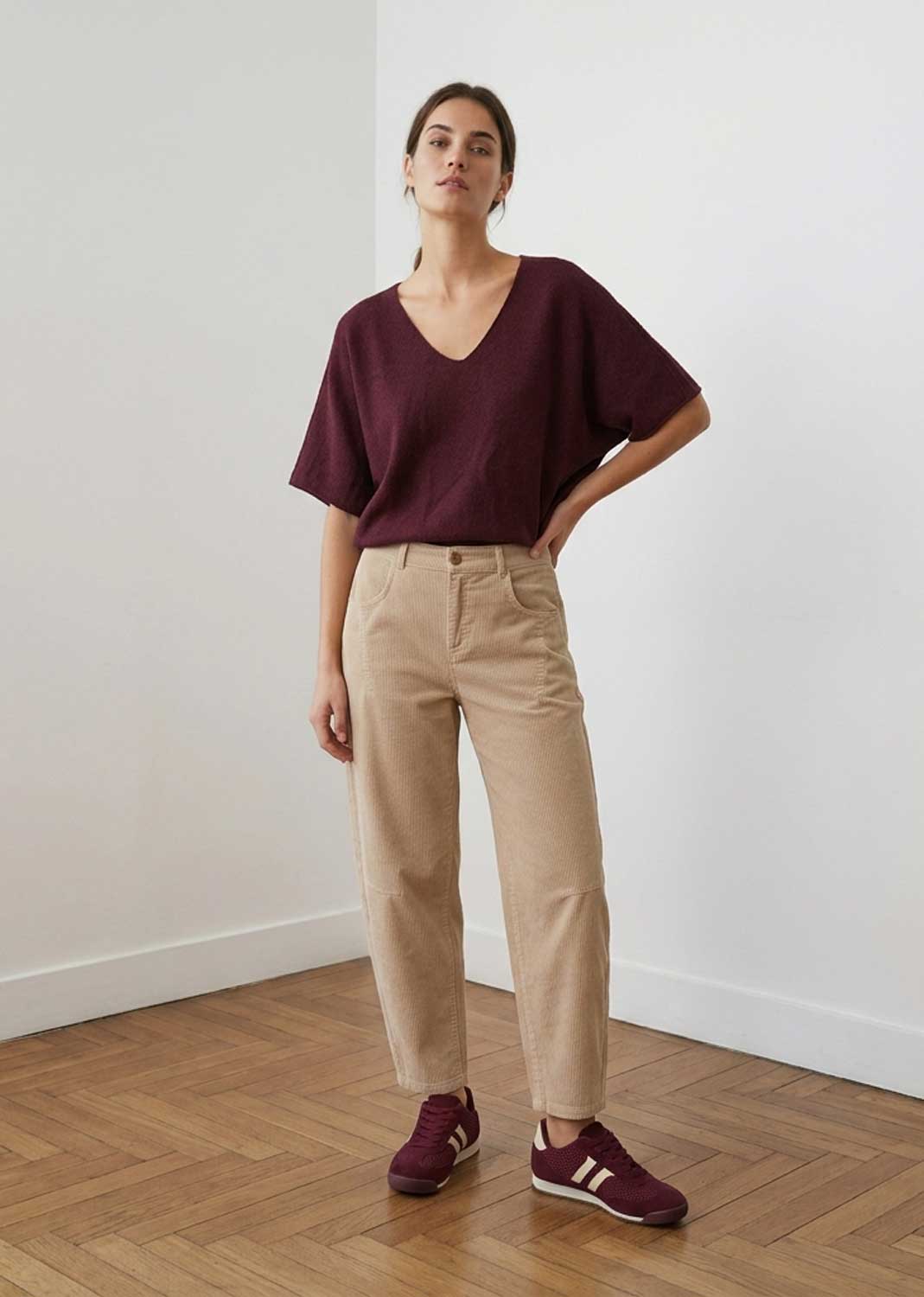 A woman stands in a minimalist room with white walls and a wooden parquet floor, wearing a loose maroon V-neck top, beige trousers, and maroon sneakers.