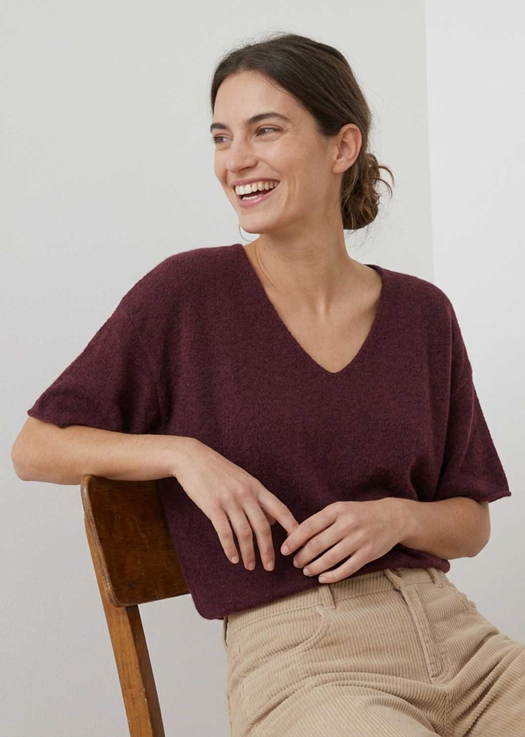 A woman smiling while wearing a short-sleeve V-neck knit top in deep burgundy, paired with beige corduroy pants, seated on a wooden chair against a plain light background.