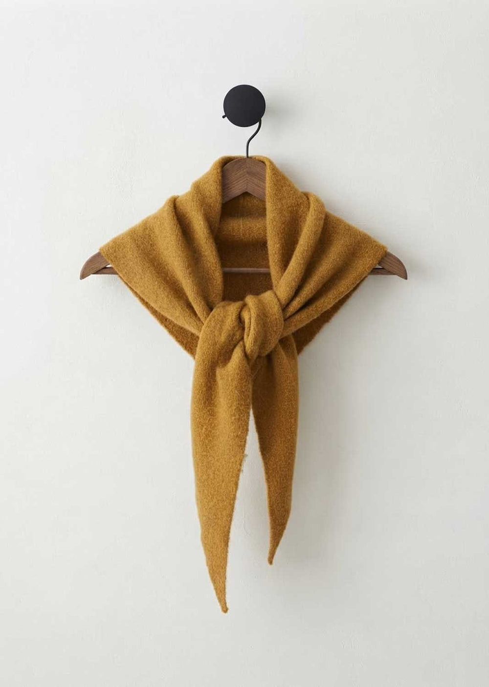 A mustard yellow knit scarf tied in a knot and draped over a wooden hanger, hanging against a plain off-white wall.