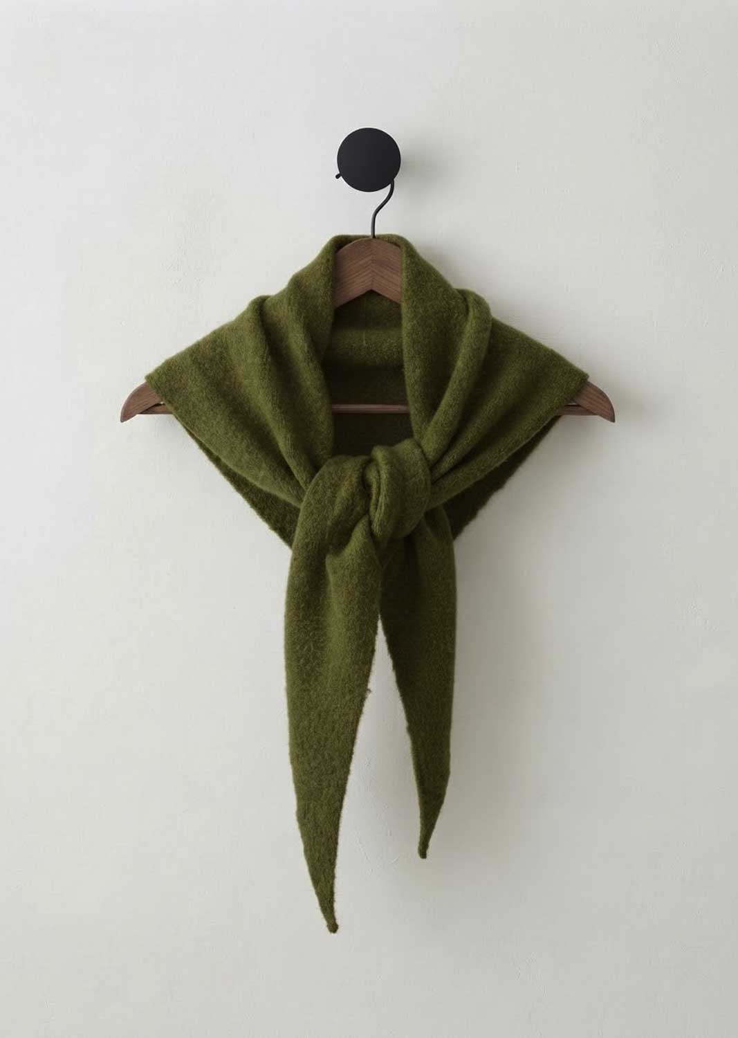 An olive green knit scarf tied in a knot and draped over a wooden hanger, hanging from a black wall hook against a plain off-white wall.