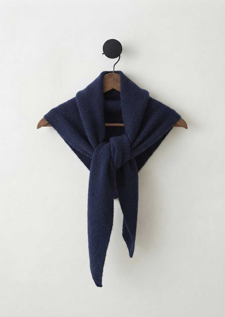 A dark navy blue knit scarf tied in a loose knot and draped over a wooden hanger, hanging against a plain off-white wall.