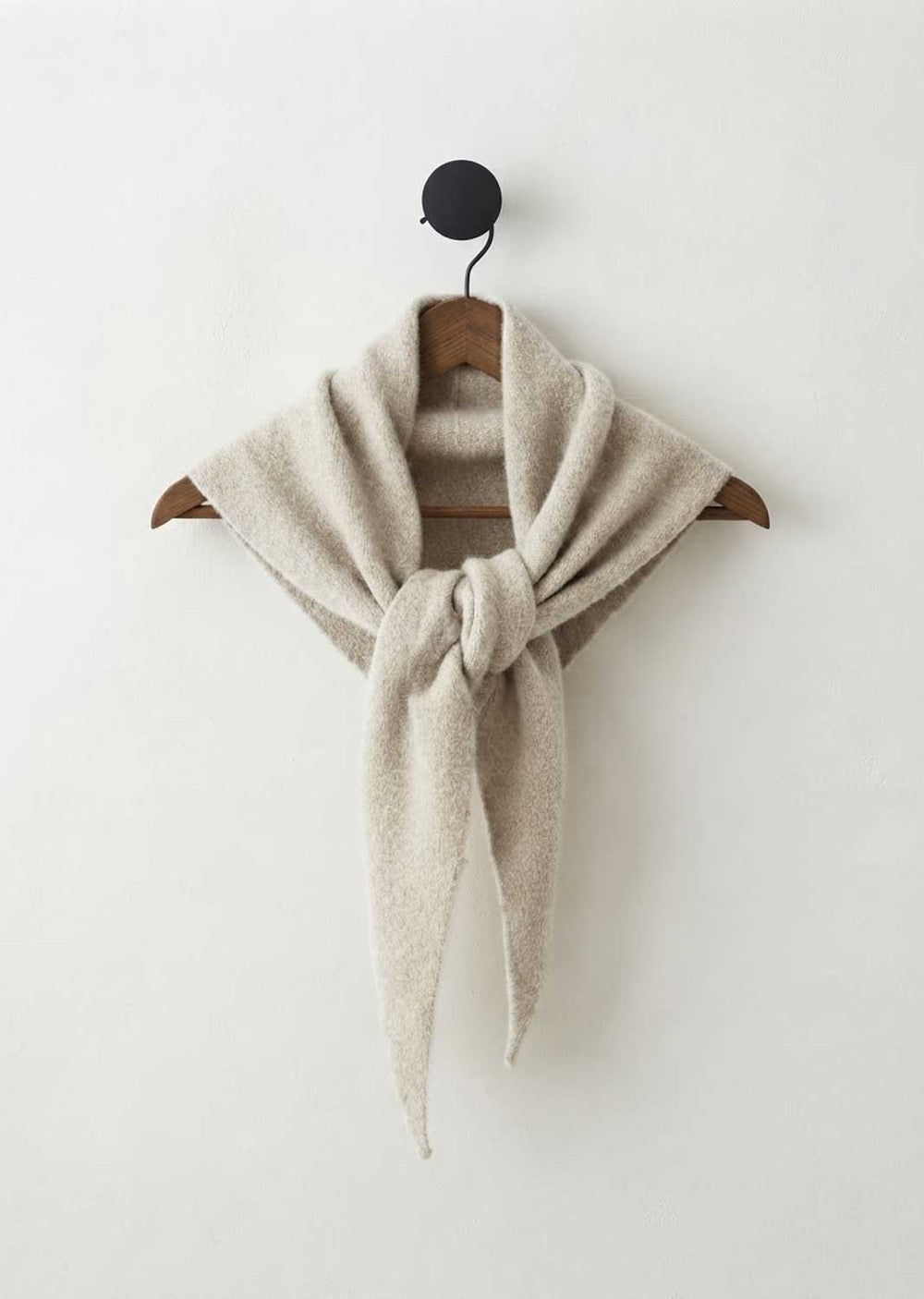 A light beige knit scarf tied in a knot and draped over a wooden hanger, hanging from a black wall hook against a plain off-white wall.