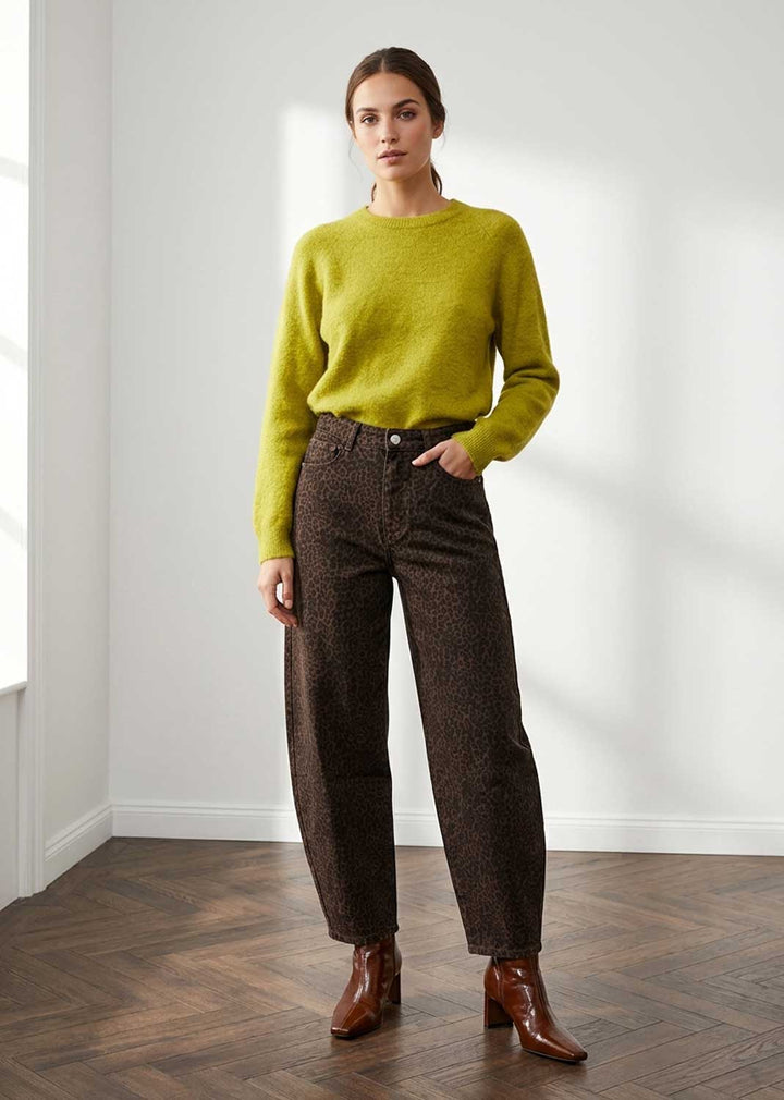 A woman stands in a bright room with white walls and a wood floor, wearing a bright yellow-green knit sweater, dark brown patterned trousers, and brown ankle boots.