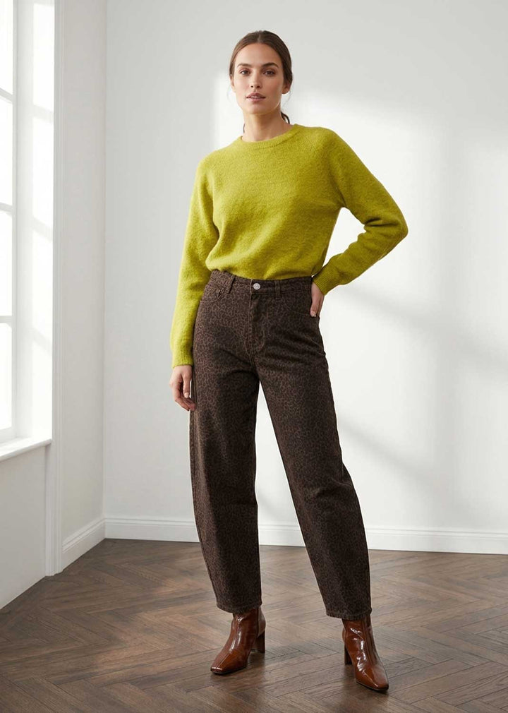A woman stands in a bright room wearing a bright yellow-green crewneck sweater, dark brown barrel leg jeans, and brown leather ankle boots.