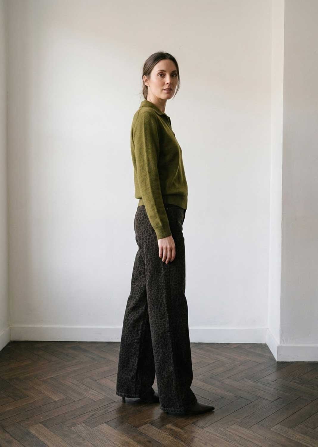 A woman stands in profile in a minimalist room with white walls and a dark wood herringbone floor, wearing an olive green knit mock-neck sweater and dark textured wide-leg trousers.