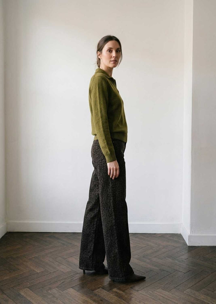A woman stands in profile in a minimalist room with white walls and a dark wood herringbone floor, wearing an olive green knit mock-neck sweater and dark textured wide-leg trousers.