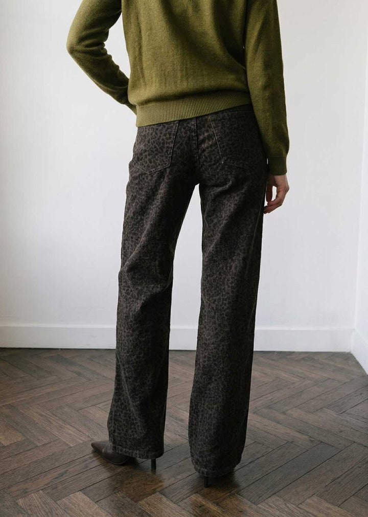 A person shown from the back wearing dark patterned wide-leg trousers and an olive green sweater, standing on a wood herringbone floor against a white wall.