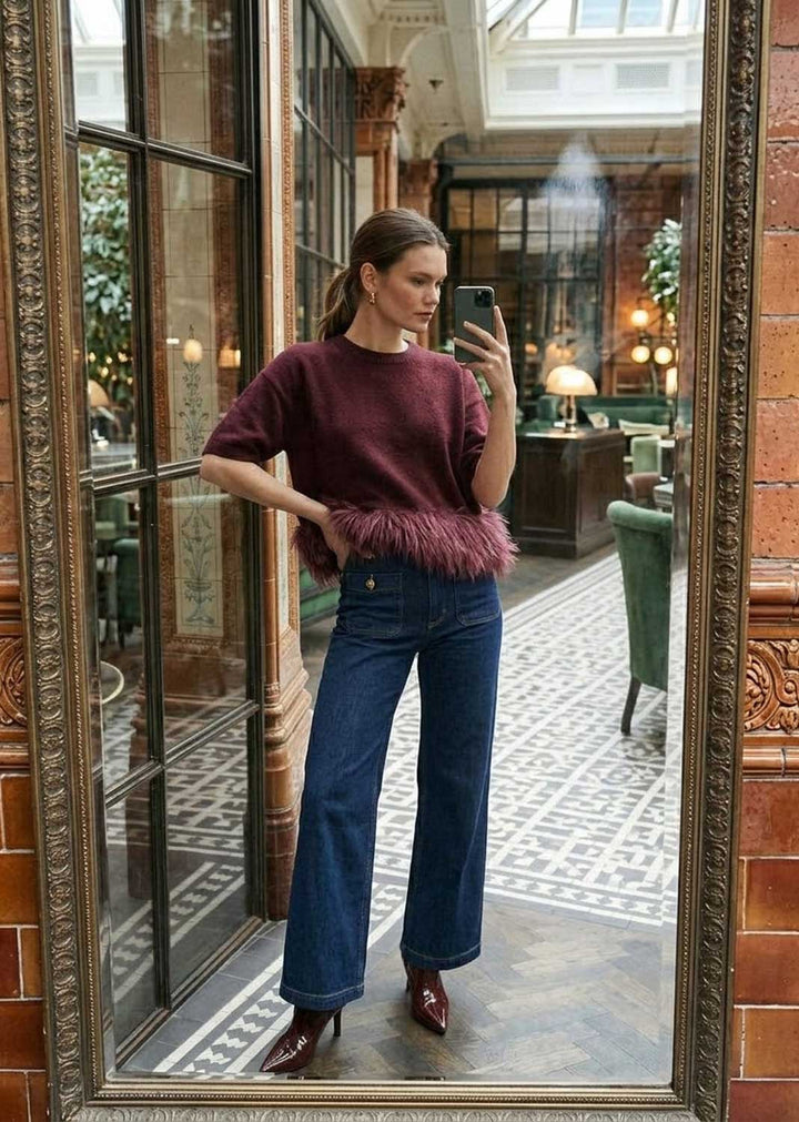 A woman takes a mirror selfie in a grand, ornate indoor space, wearing a burgundy short-sleeve sweater with a fluffy feather-trim hem and dark blue wide-leg jeans.