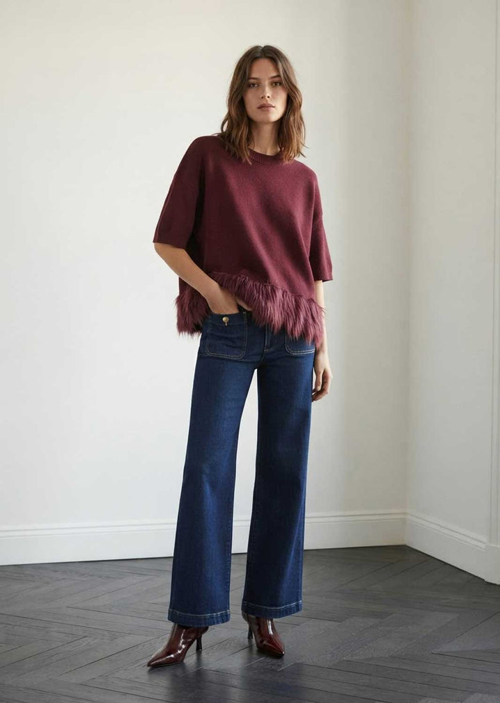 A woman stands in a minimalist room with white walls and dark wood flooring, wearing a short-sleeve maroon knit sweater with a fringe hem, dark blue wide-leg jeans, and brown leather boots.