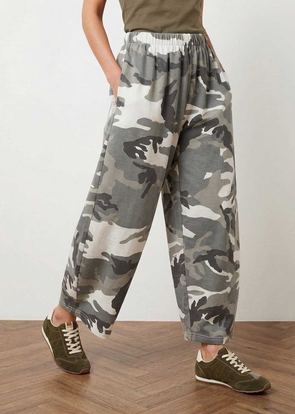 A person wearing loose, wide-leg jogger-style pants with a gray, black, and off-white camouflage pattern, paired with olive green sneakers and a matching olive top, standing on a wood floor against a plain light wall.